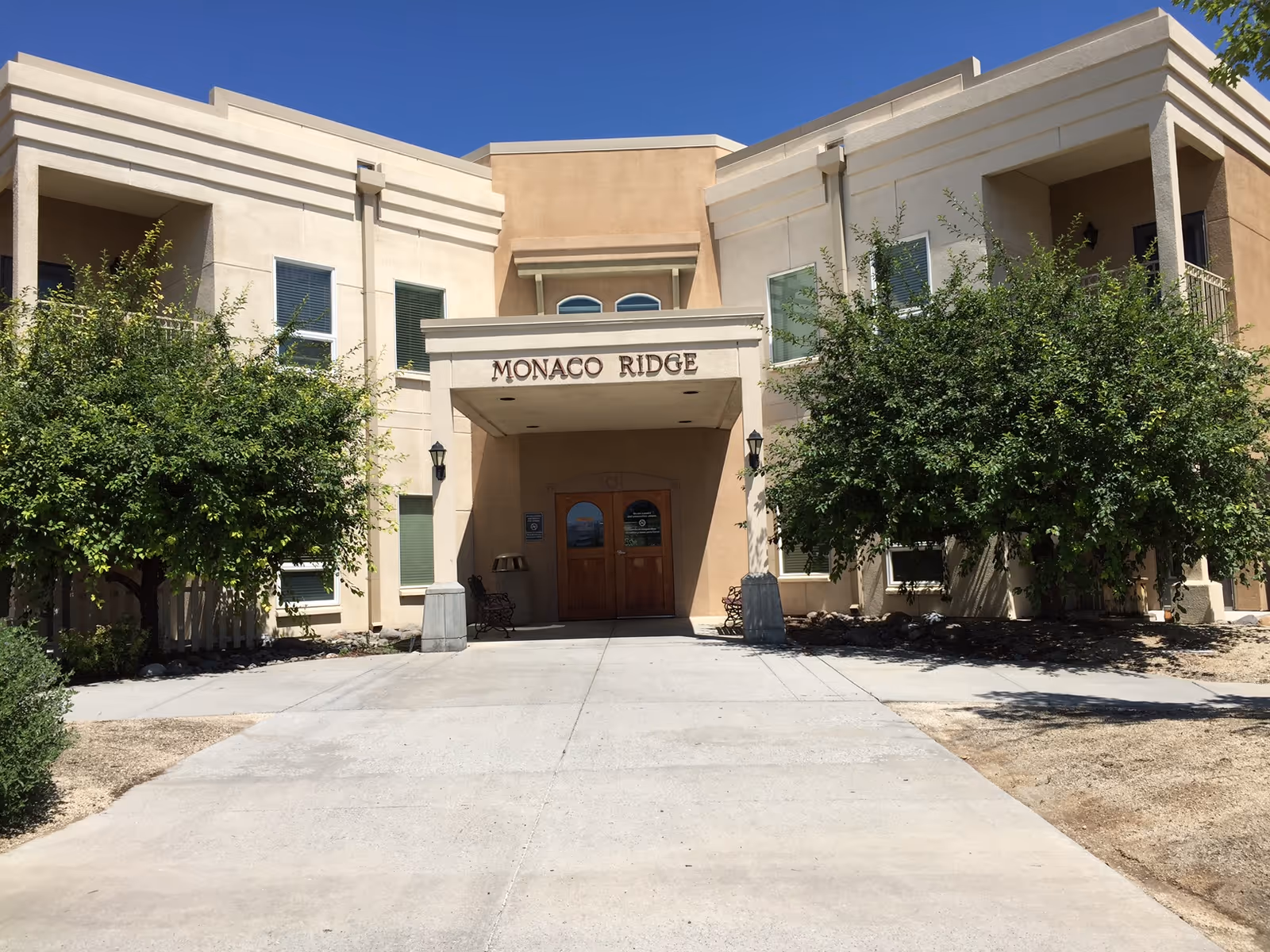 Front entrance of the Monaco Ridge building showing the canopy with 'MONACO RIDGE' signage, double wooden doors, and trees on either side.