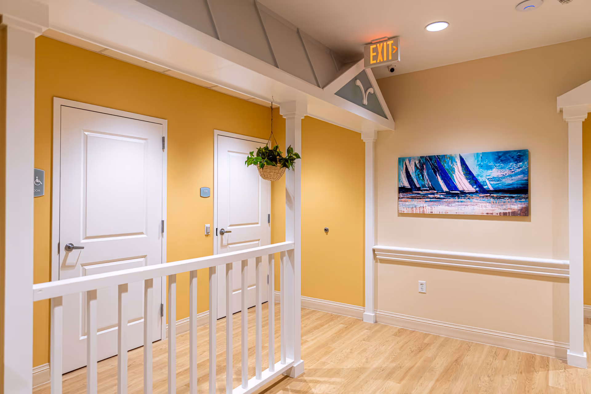 Bright interior hallway with yellow walls, white doors and railing, a hanging plant, exit sign, and a colorful sailboat painting.
