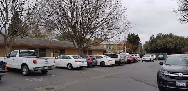 Parking lot in front of a single-story care facility with several parked cars and leafless trees under an overcast sky.