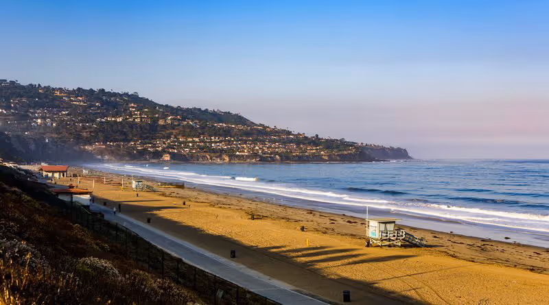 Sunny coastal beach with boardwalk, lifeguard hut, and hillside houses along shore