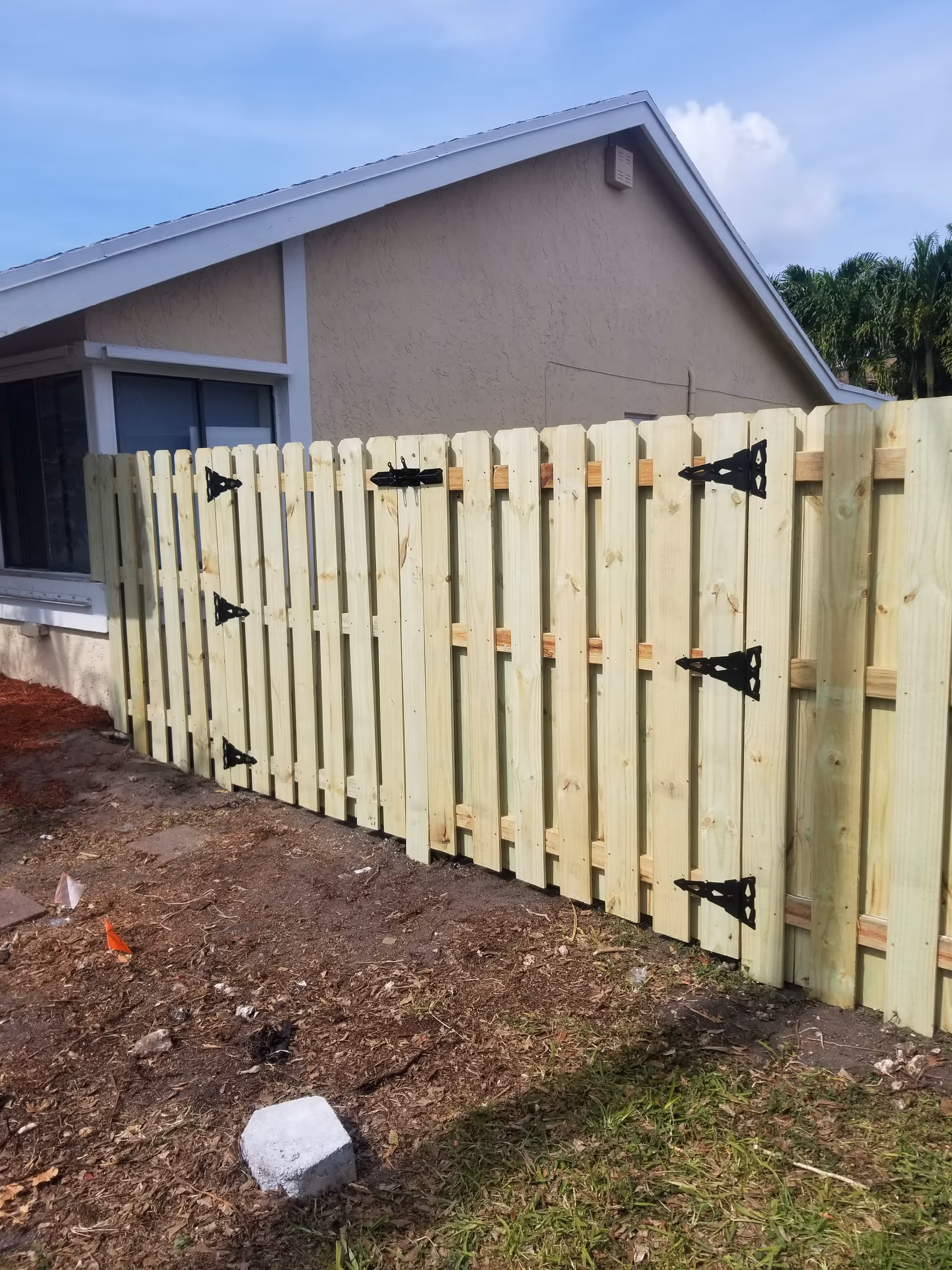 Light wooden fence runs along a beige building with dirt ground and debris nearby.