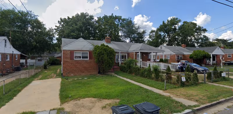 A single-story brick residential building with a gray roof and a small front porch with a white railing. There is a green lawn in front with a concrete driveway on the left side and a sidewalk leading to the porch. Several trees and bushes surround the house, and neighboring similar houses are visible on either side. The sky is partly cloudy with blue patches.
