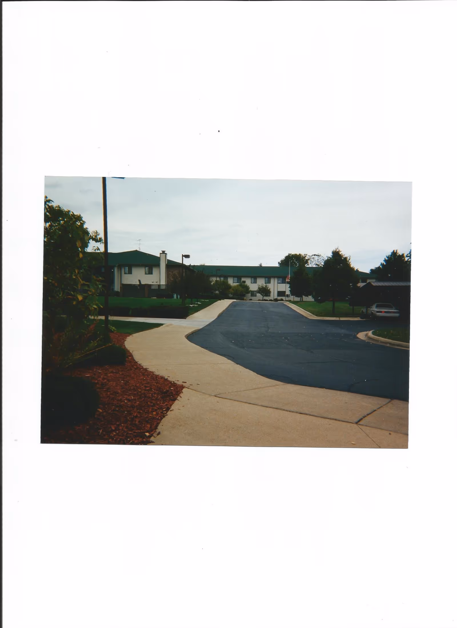 Driveway and sidewalk leading to a two-story building with a green roof, lawns and trees on either side and a parked car to the right.