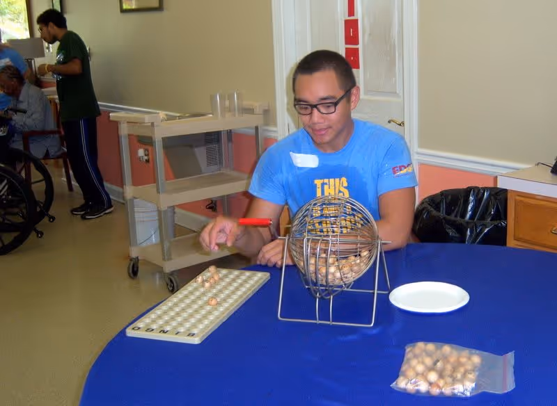 A young man wearing glasses and a blue shirt is sitting at a table covered with a blue tablecloth, playing bingo using a bingo cage and a bingo card. In the background, there are other people, including one in a wheelchair, and a utility cart against the wall in a room with beige walls and a window.