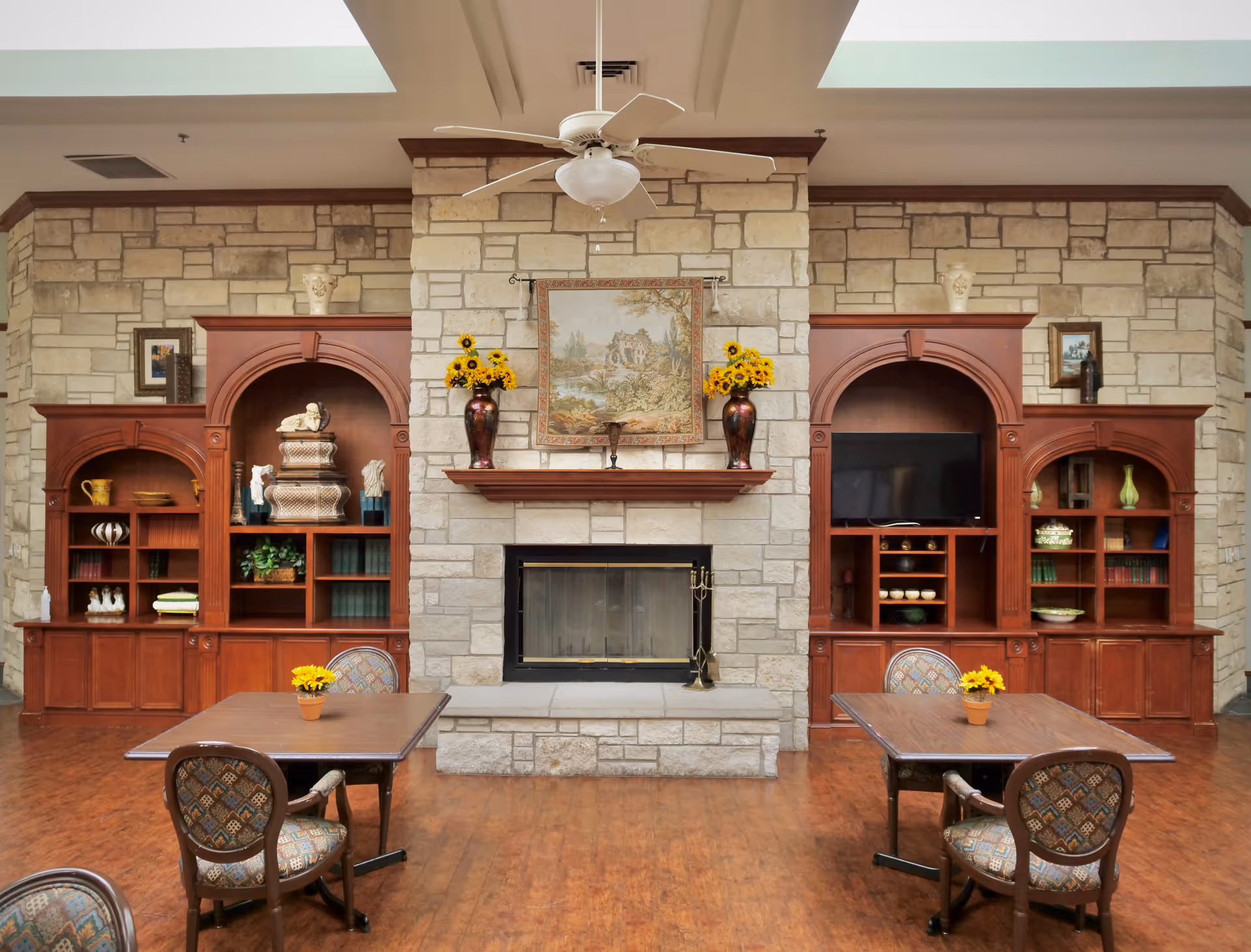 A cozy common area with a stone fireplace in the center, flanked by wooden built-in shelves and cabinets. The shelves contain decorative items, books, and a TV. Two tables with patterned chairs and small flower pots are placed in front of the fireplace. A ceiling fan hangs above, and the room has a warm, inviting atmosphere.