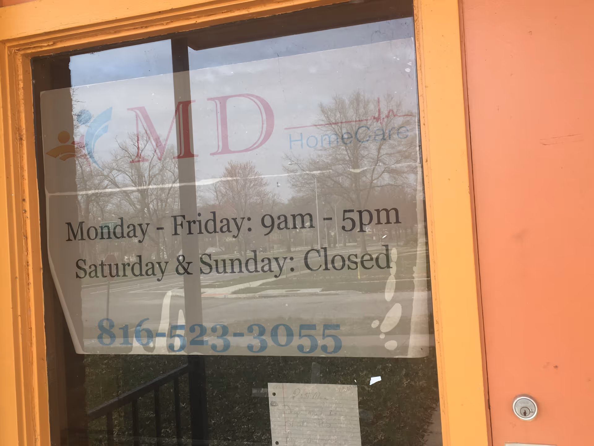 Glass door displaying MD HomeCare sign and hours, orange frame, outdoor scene beyond.