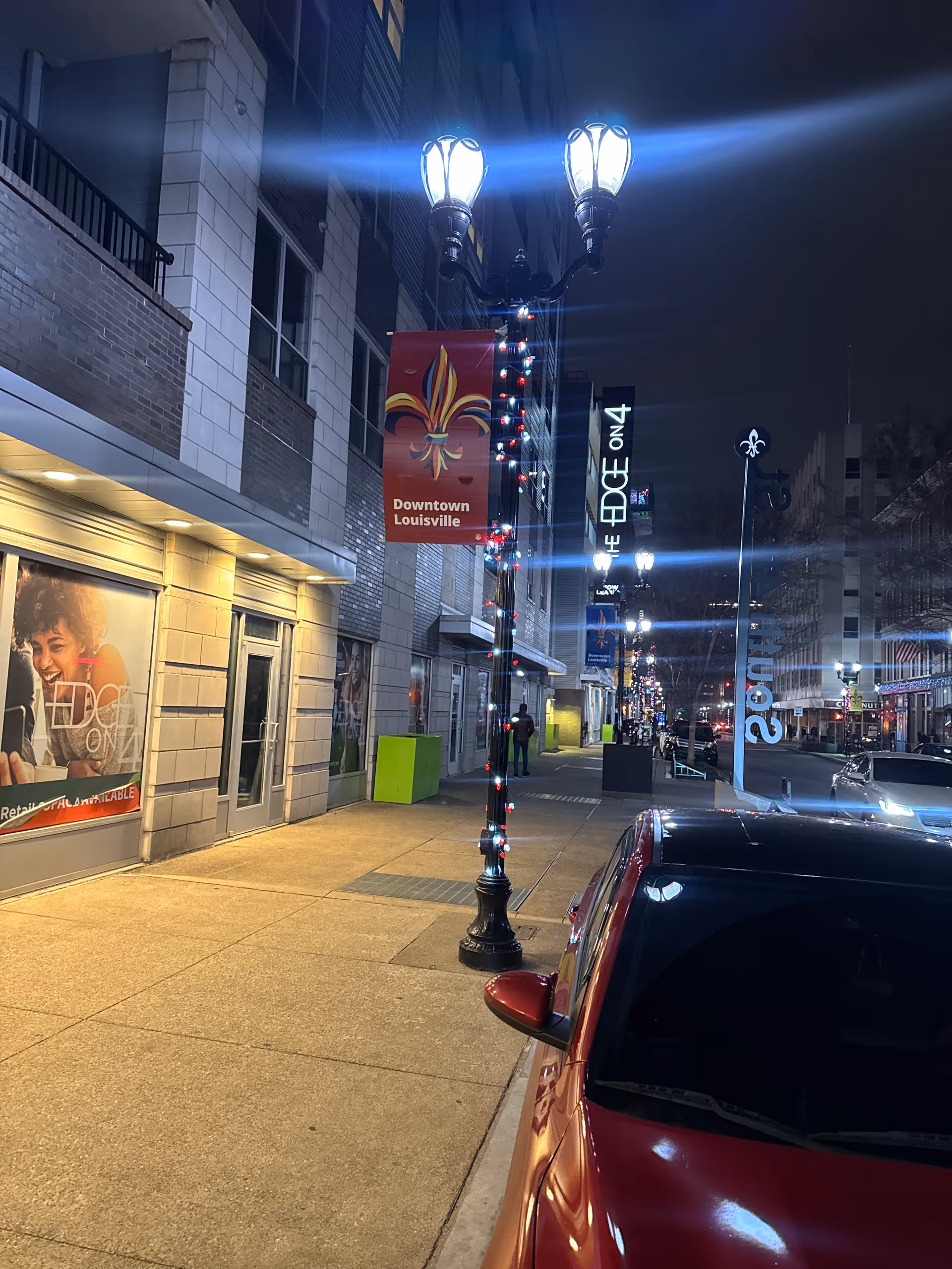 Nighttime downtown Louisville street with lit banners, storefronts, and a red car along the curb