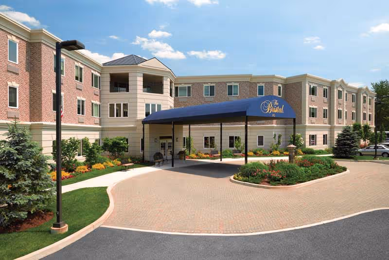 Front entrance of The Bristal assisted living building with a blue canopy over the driveway, landscaped beds, and a three-story brick façade.