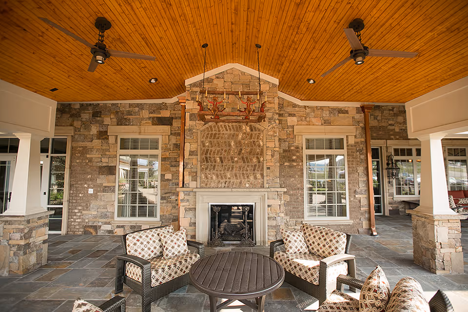 Covered stone patio with a central fireplace, wicker seating and a wooden ceiling with fans.