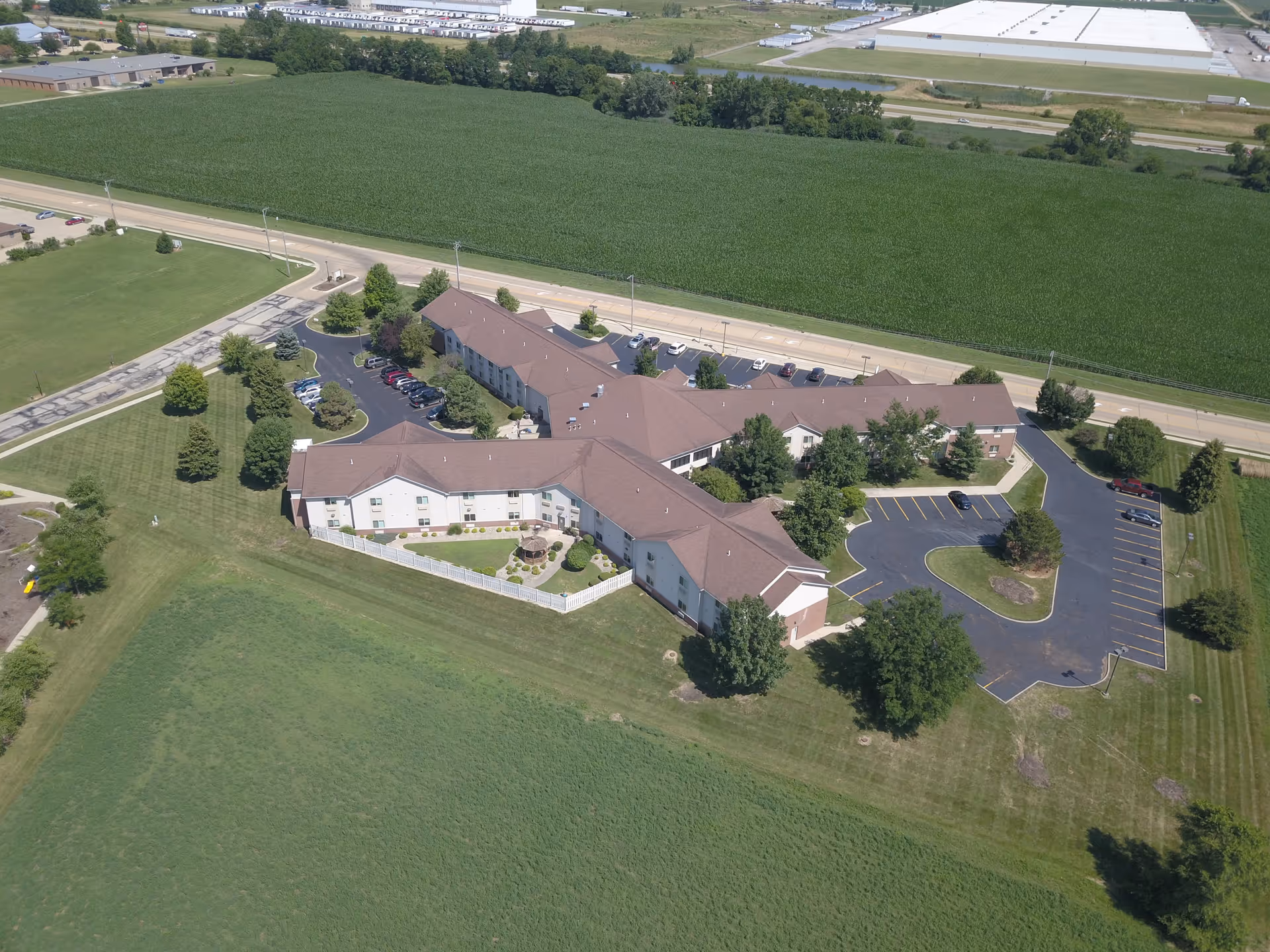 Aerial view of a large senior living facility building with brown roofs surrounded by green lawns and trees. The facility has multiple parking lots with several cars parked. The building is adjacent to a road and large agricultural fields.