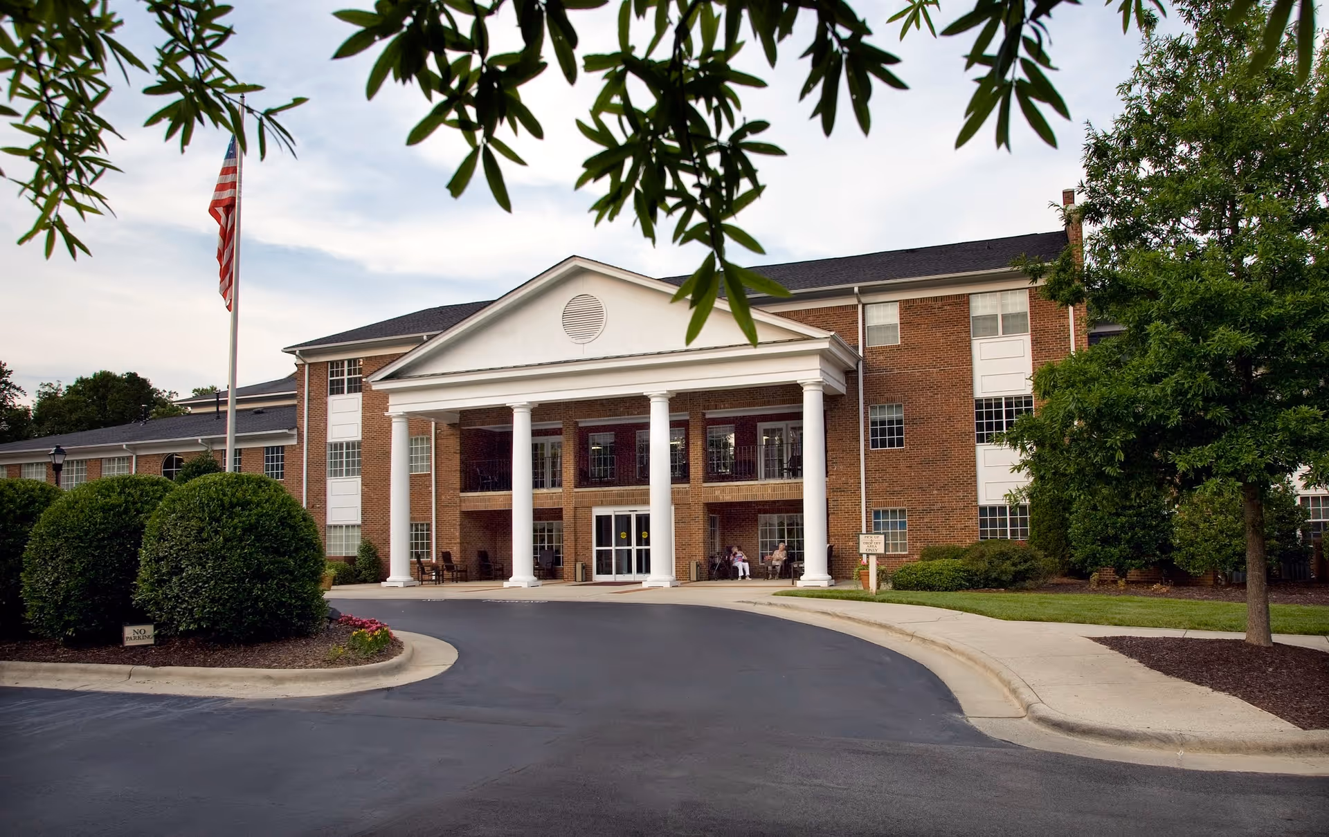 Front exterior view of a large brick building with white columns at the entrance, a curved driveway, neatly trimmed bushes, an American flag on a flagpole, and trees surrounding the area.
