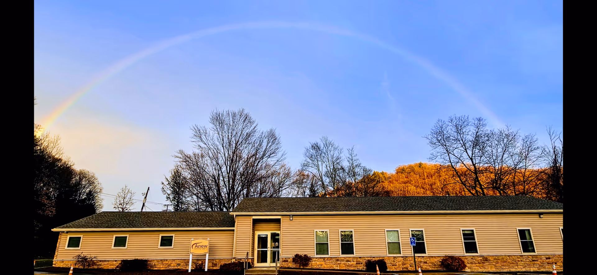 Single-story beige facility with Anew sign, parking area, and a rainbow in the sky.