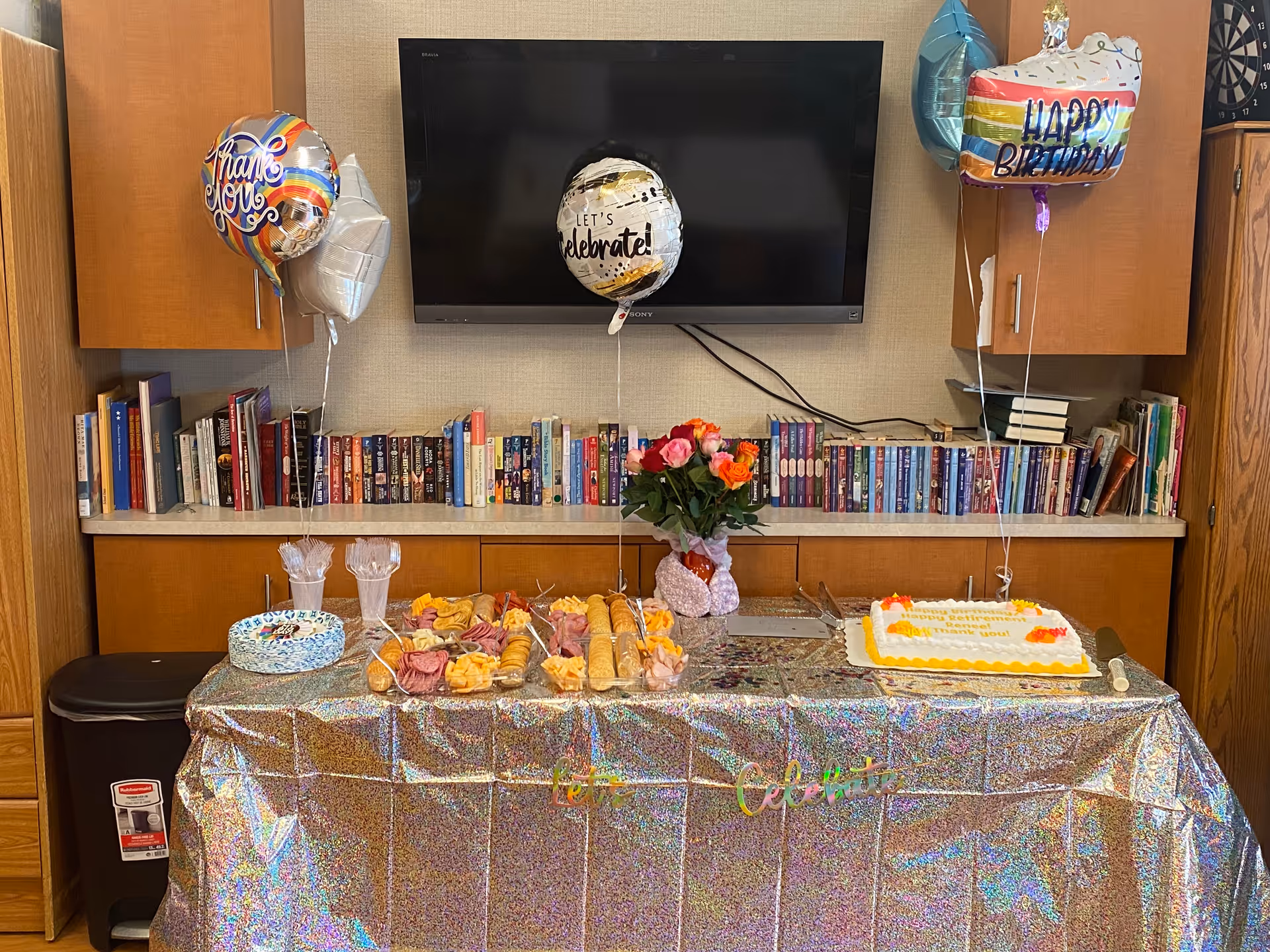 Birthday celebration setup with cake, pastries, balloons, and flowers on a book-lined wall.