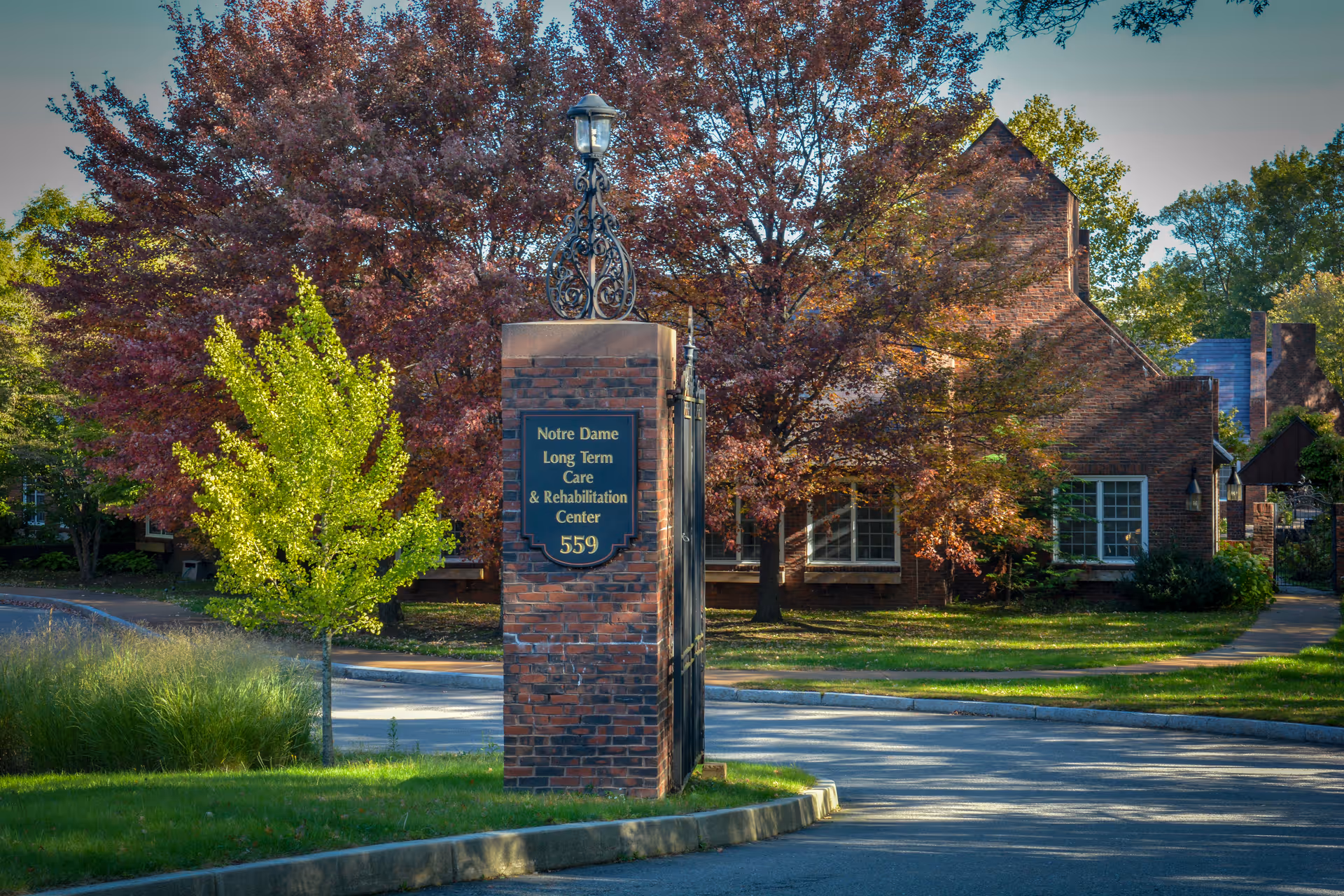 Entrance sign for Notre Dame Long Term Care & Rehabilitation Center with the number 559, surrounded by trees with autumn foliage and a brick building in the background.