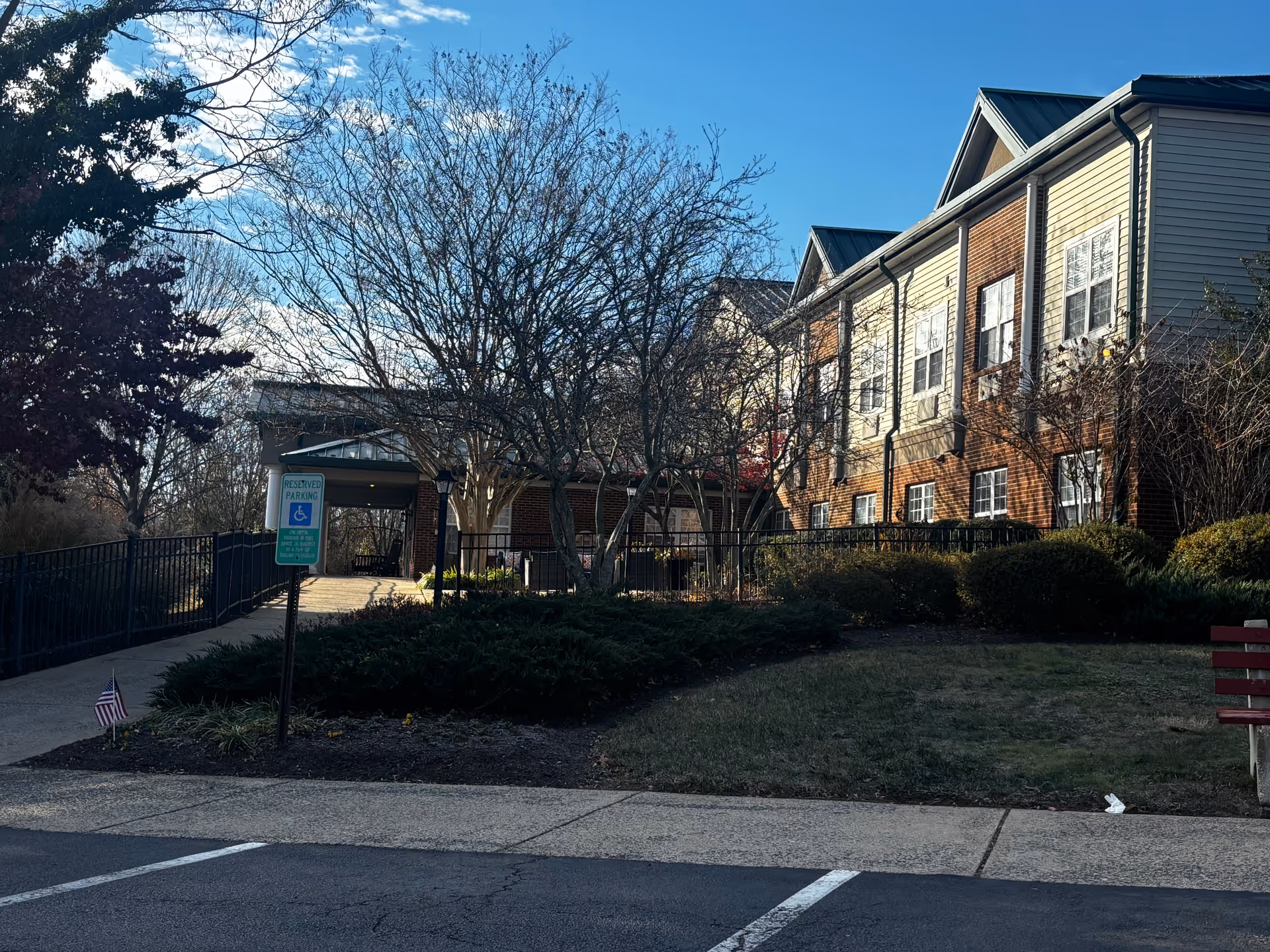 Exterior view of a senior living facility building with a brick and siding facade, multiple windows, and a metal roof. There is a paved walkway with a handrail leading to the entrance, a reserved parking sign for handicapped parking, some bushes and trees without leaves, and a clear blue sky.