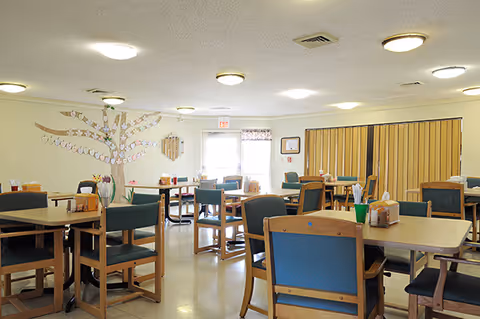 A well-lit dining room with multiple wooden tables and chairs arranged neatly. The walls are decorated with a paper tree and other crafts. The room has ceiling lights and a door with an exit sign above it.