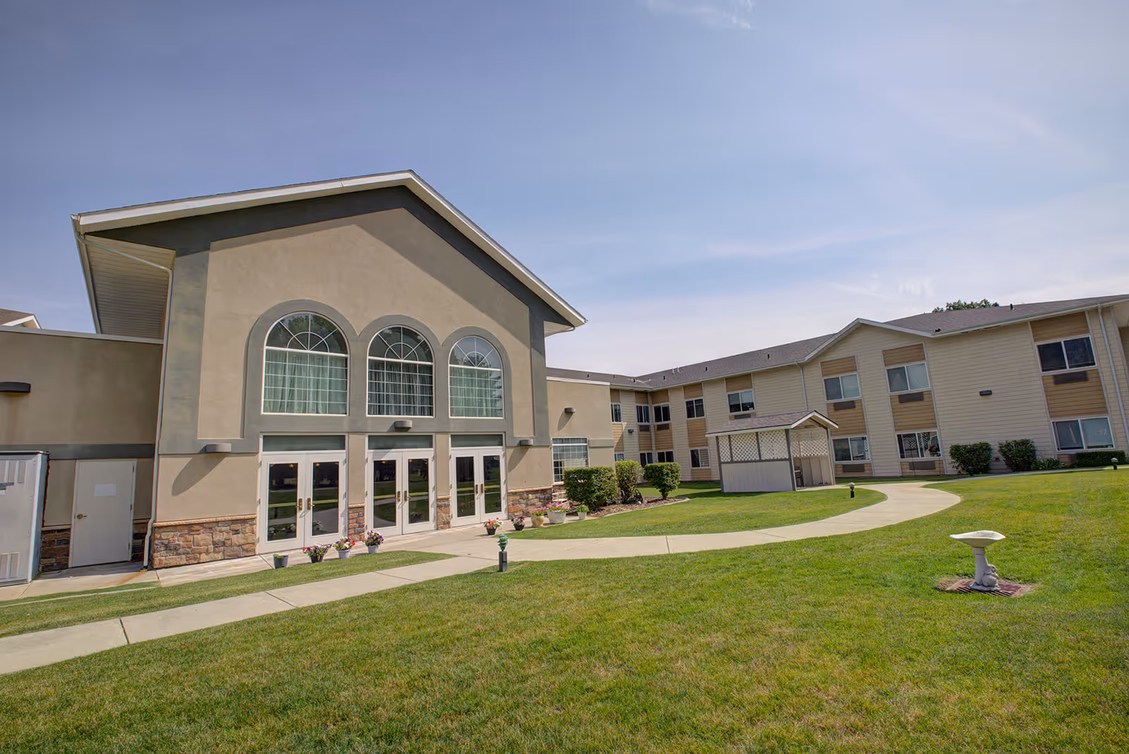 Exterior view of a senior living facility building with beige walls, large arched windows, multiple glass doors, and a well-maintained green lawn with a curved concrete pathway under a clear blue sky.