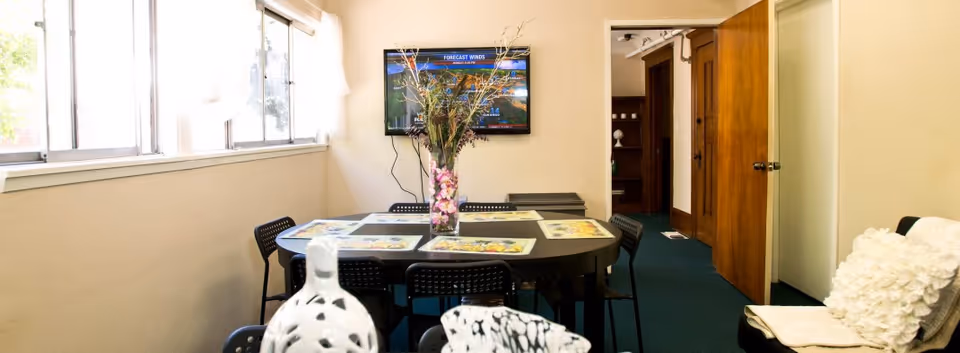 A dining area with a rectangular black table surrounded by black chairs. The table is set with placemats and a vase with flowers in the center. A flat-screen TV is mounted on the wall showing a weather forecast. There are windows on the left side letting in natural light, and an open wooden door leads to another room. A cushioned chair with a white pillow is visible on the right side.
