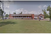 Front view of a single-story brick nursing home with a porch, chairs, and a grassy lawn.