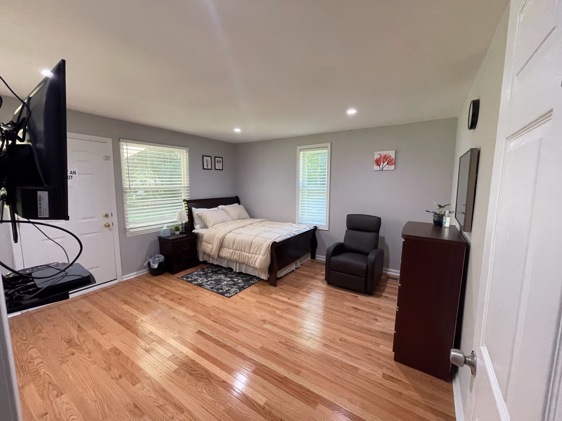 A clean and simple bedroom with light gray walls and wooden flooring. The room features a bed with white bedding, a nightstand with a lamp and tissue box, a black recliner chair, a dark wooden dresser with a plant and candle, two windows with blinds, and a wall-mounted TV. There are a few framed pictures on the walls.