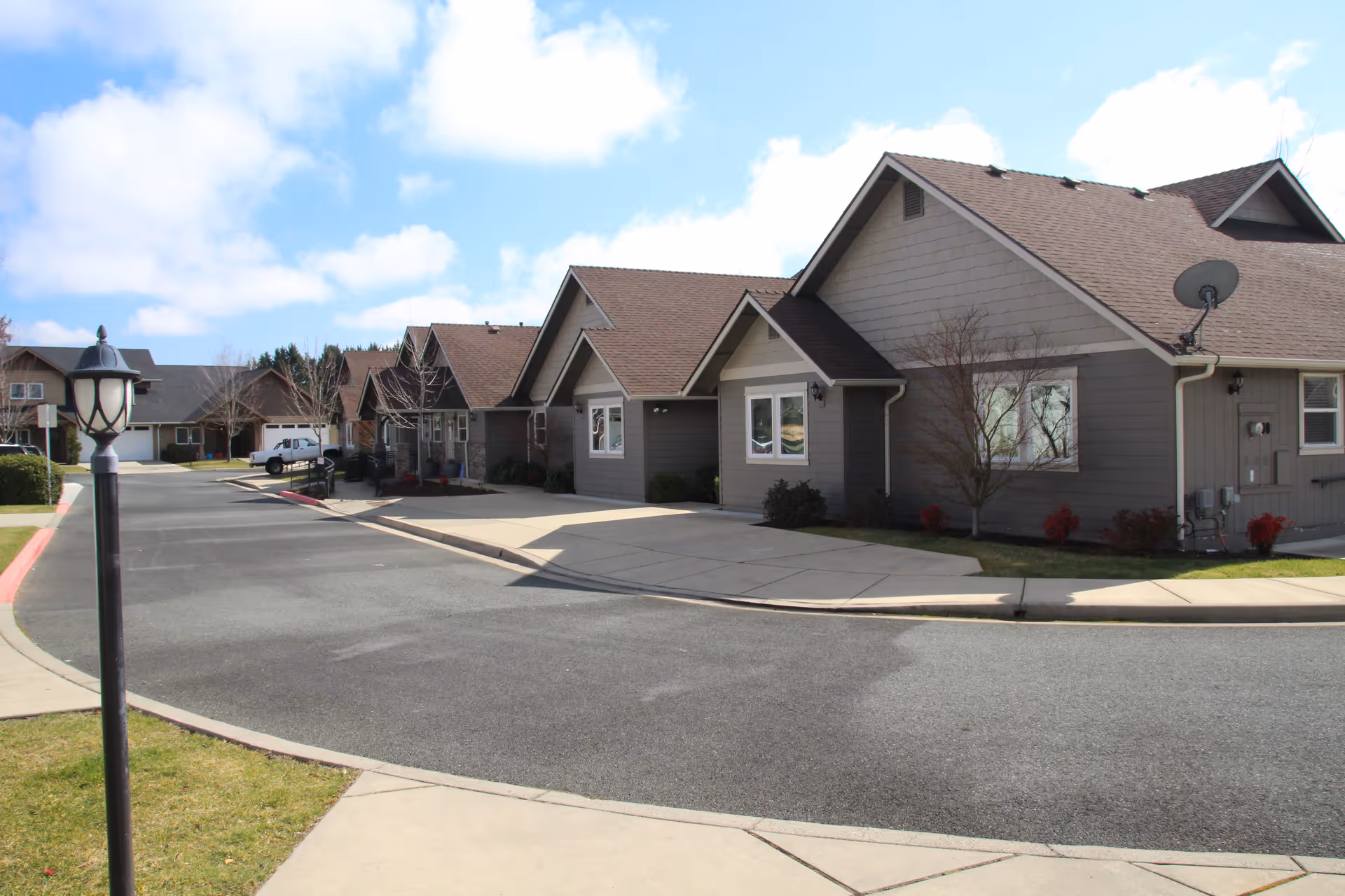 A row of single-story residential buildings with gray siding and brown shingled roofs under a partly cloudy blue sky. There is a paved road and sidewalk in front, along with a black street lamp on the left side of the image. Some small trees and shrubs are planted near the buildings.
