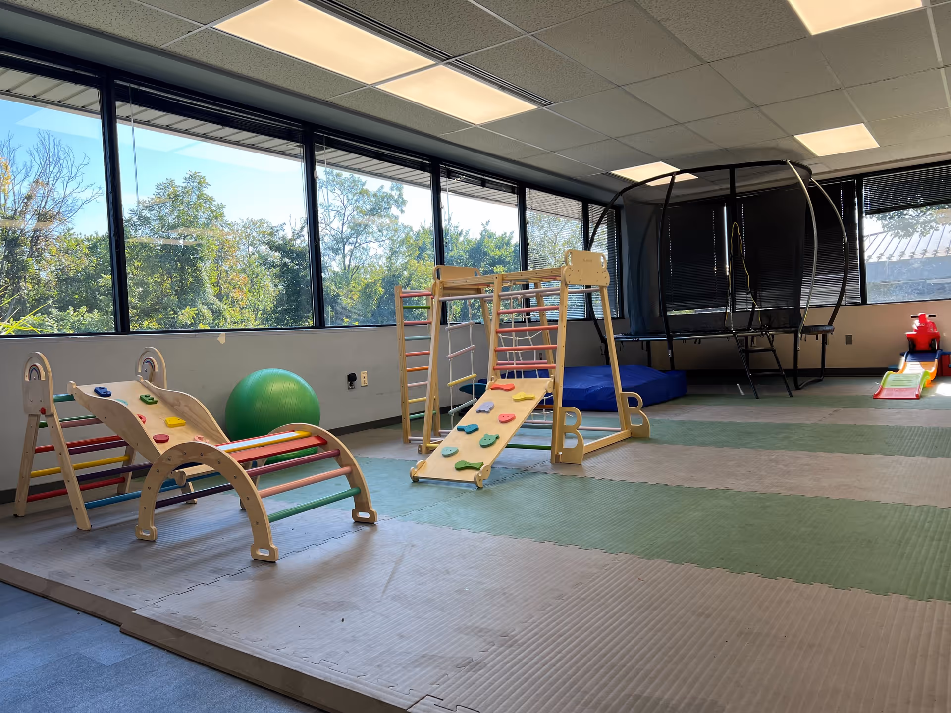 Indoor playground area with wooden climbing frames, mats, and a green exercise ball by windows.