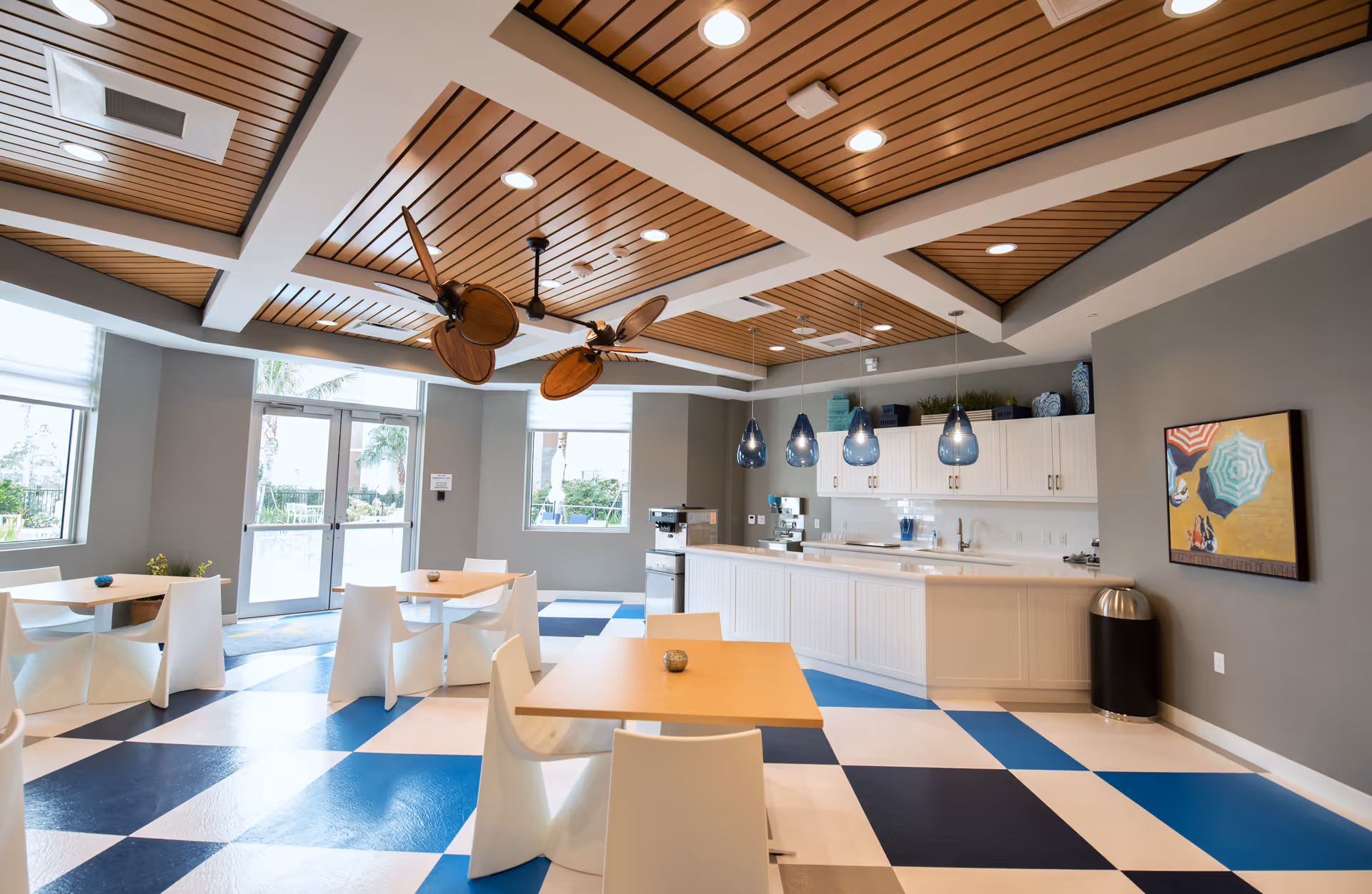 Bright and modern dining area with white tables and chairs, a checkered blue and white floor, a wooden slatted ceiling with ceiling fans, pendant lights above a white kitchen counter, large windows, and a door leading outside. A colorful painting is mounted on the gray wall.