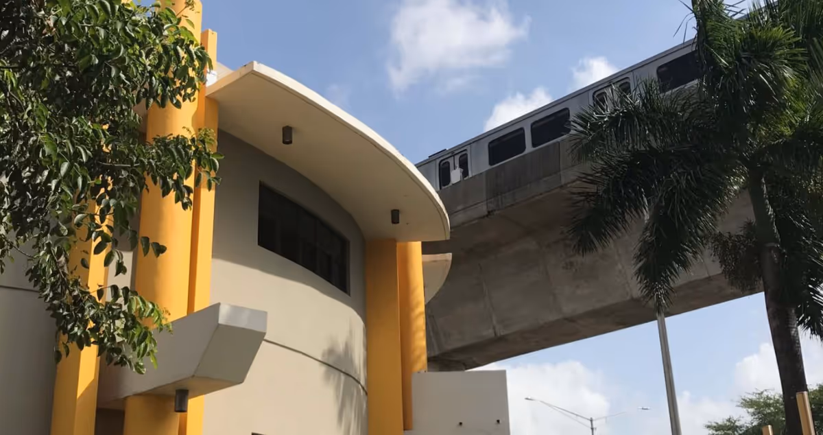 Curved, yellow modern building facade with tall columns, palm trees, and an elevated train track.