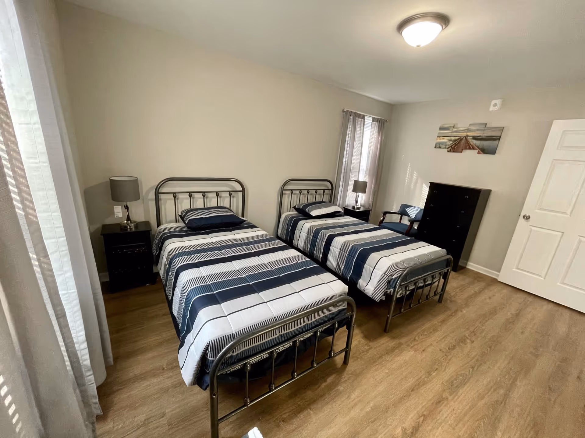 A bedroom with two single metal frame beds, each with striped bedding in shades of blue, white, and gray. There are two nightstands with lamps on either side of the beds. A window with sheer curtains is on the left wall, and another window with blinds and curtains is on the far wall. A black dresser and a blue chair are positioned against the far wall, with a wall painting above the dresser. The room has light-colored walls and wood flooring.