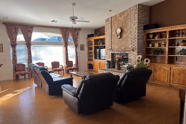 Spacious living room with leather recliners and sofas facing a brick fireplace and built-in wooden shelving, with large sunlit windows and patterned drapes.