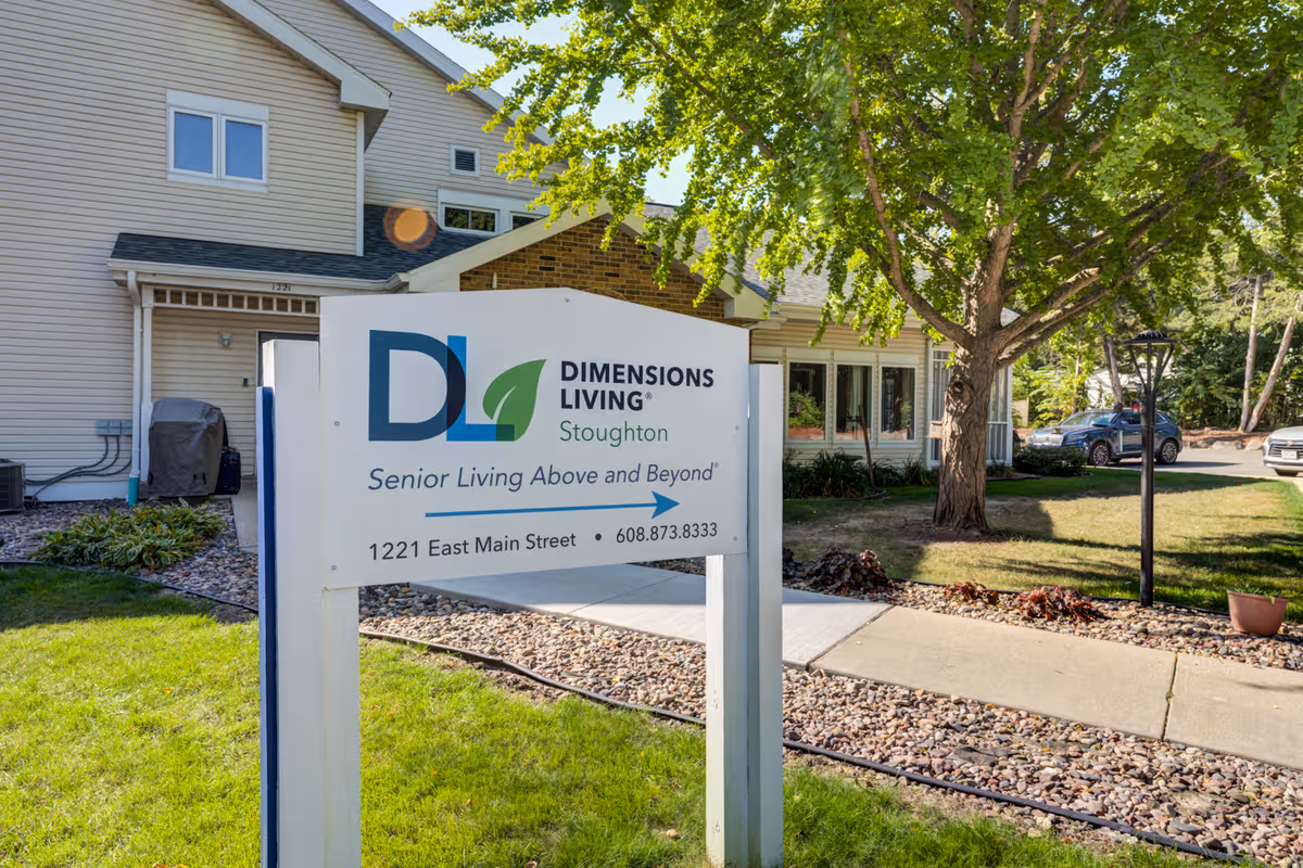 Exterior view of Dimensions Living Stoughton facility with a large front-yard sign in sunlight.