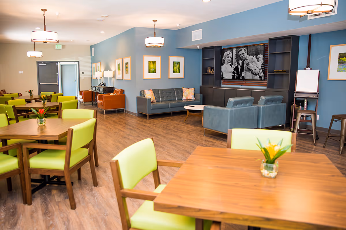 Bright senior living lounge with green chairs, wooden tables, and blue accent walls.