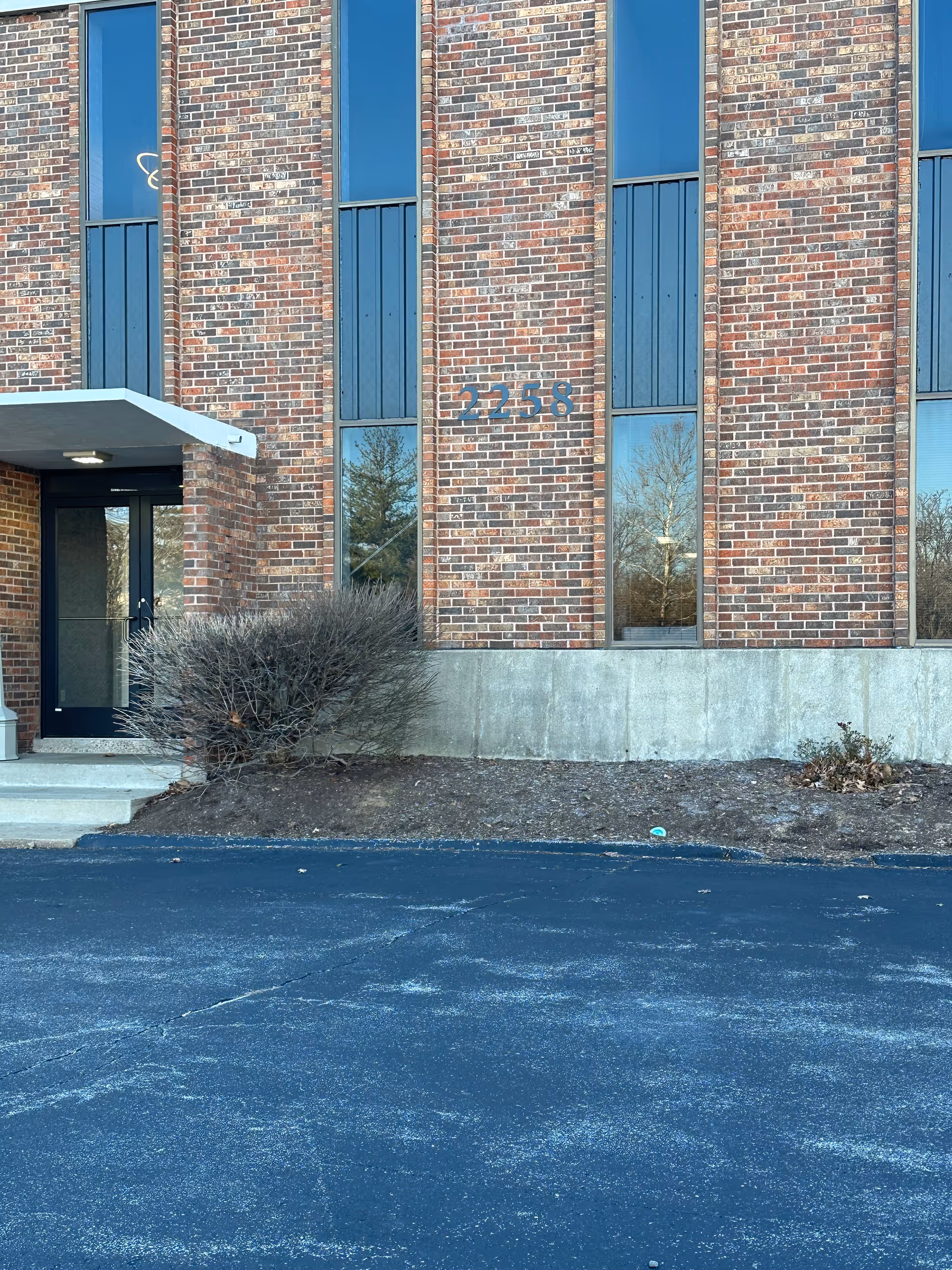 Brick building exterior with entrance, tall windows, and a paved front parking area.