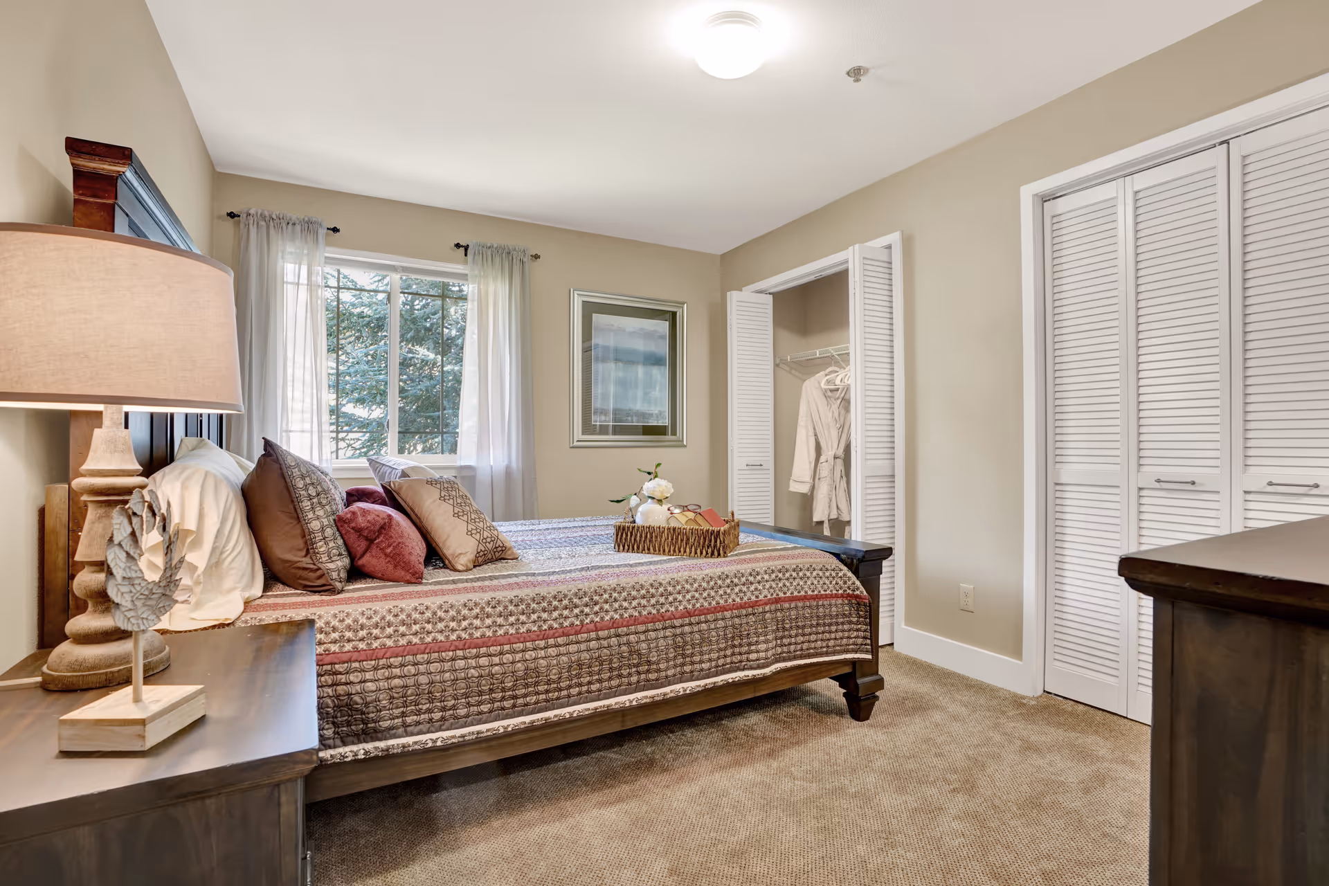 A cozy bedroom with a large bed covered in a patterned quilt and multiple pillows. There is a wooden nightstand with a lamp and decorative pineapple on the left side of the bed. A window with sheer white curtains lets in natural light. On the right side, there is an open closet with white louvered doors and a robe hanging inside. The walls are painted beige and the carpet is light brown.