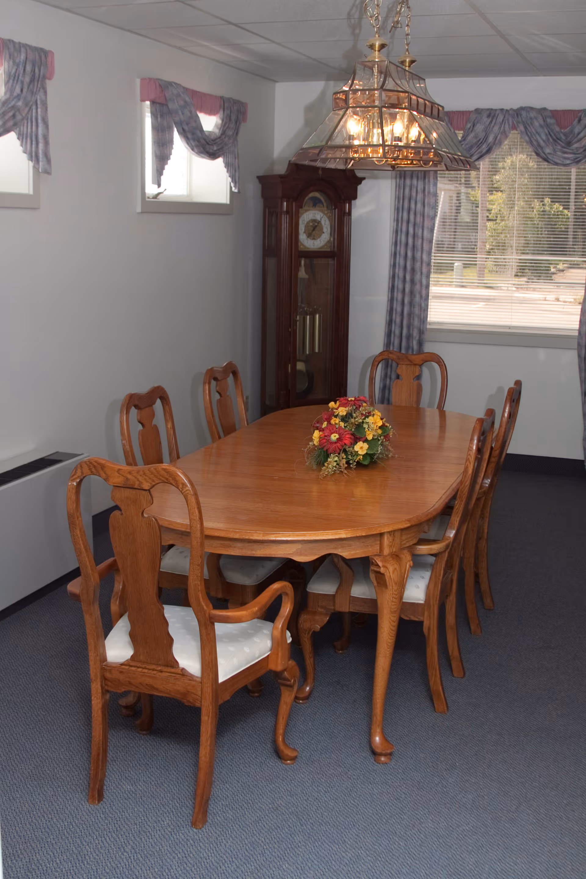 A dining room with a wooden oval dining table surrounded by six wooden chairs with white cushioned seats. A floral centerpiece is placed on the table. The room has carpeted flooring, two small windows with blue and pink valances, a larger window with matching curtains, and a tall grandfather clock in the corner. A brass and glass chandelier hangs above the table.