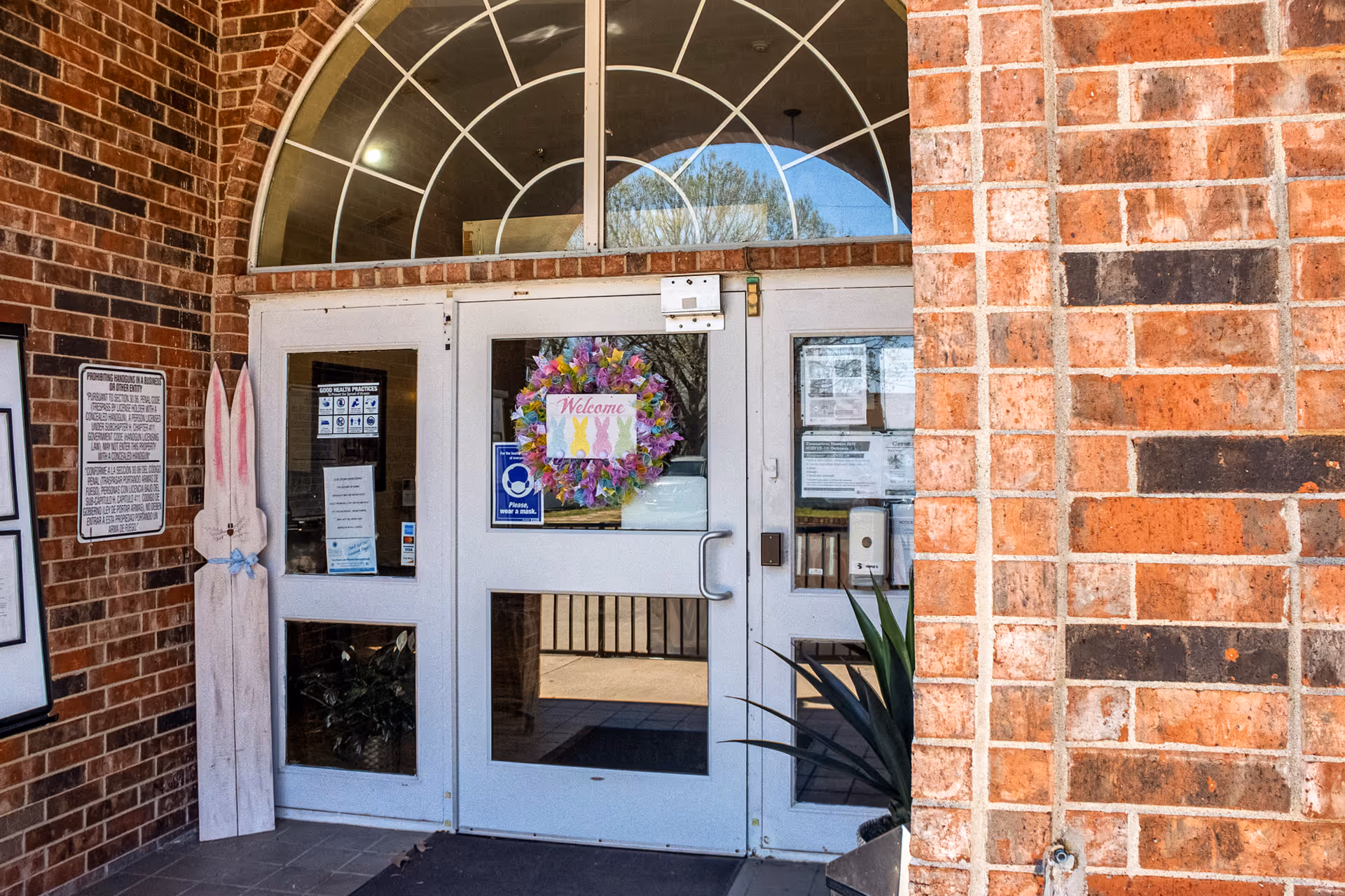 Brick building entrance with glass doors, welcoming wreath, and arched window above