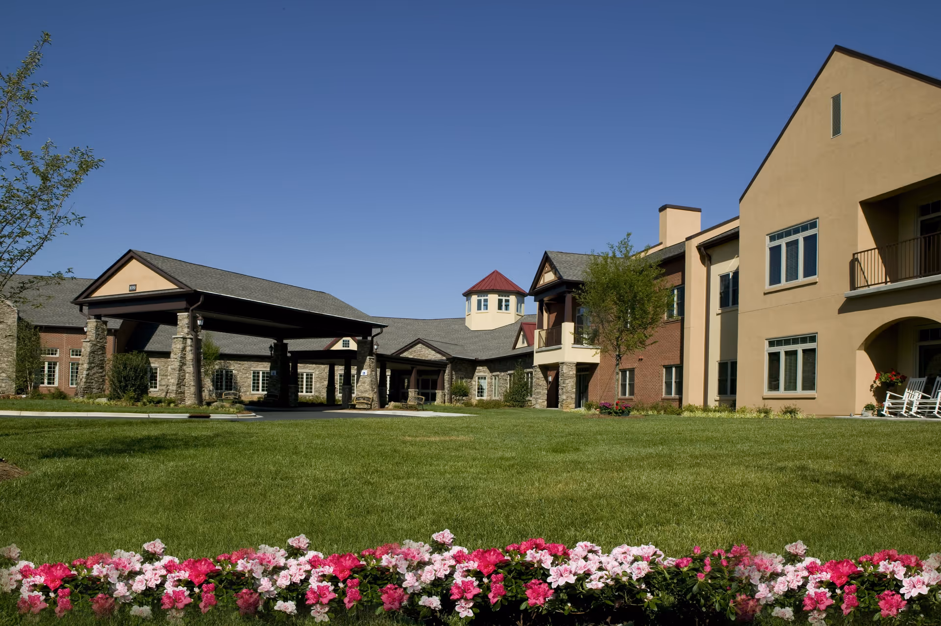 Exterior view of Pennybyrn senior living facility showing a large green lawn with a flower bed of pink and white flowers in the foreground, and a multi-section building with beige and brick walls under a clear blue sky.