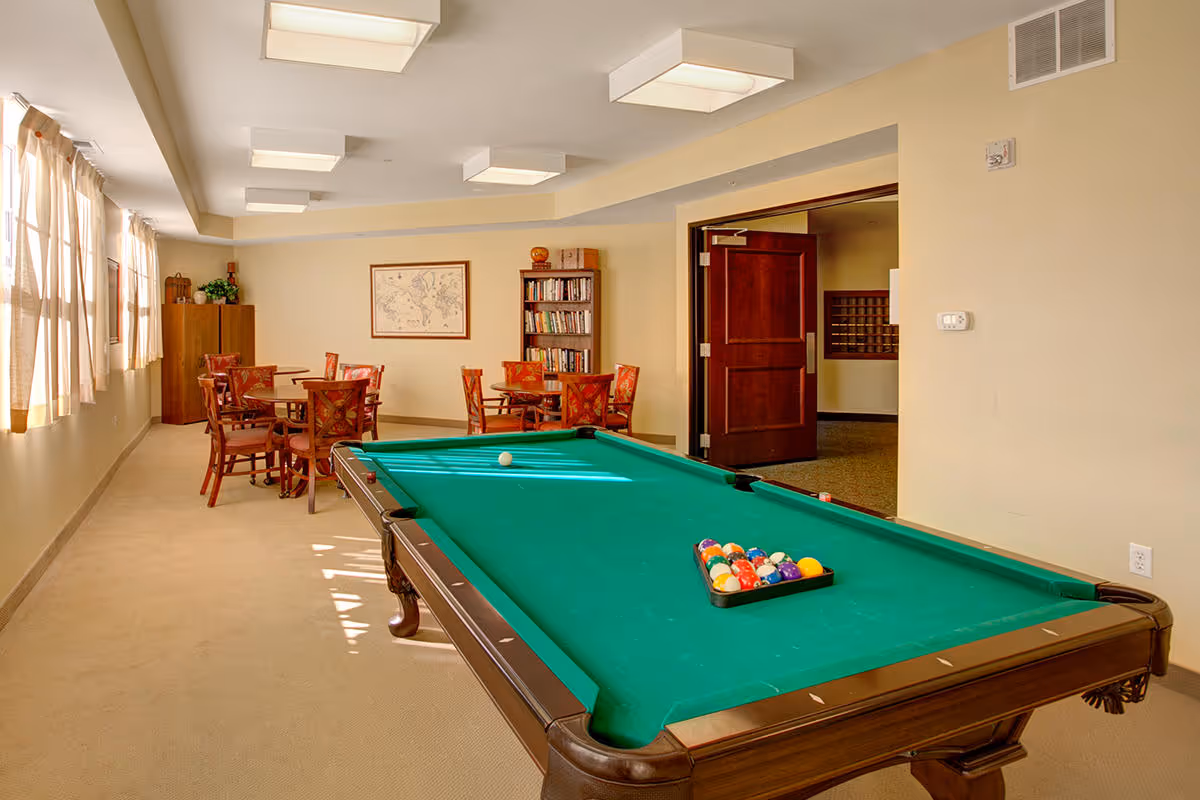 A well-lit recreational room with a green pool table in the foreground, several wooden tables and chairs in the background, a bookshelf filled with books, and a framed map on the wall. The room has beige walls, carpeted floor, and large windows with curtains letting in natural light.