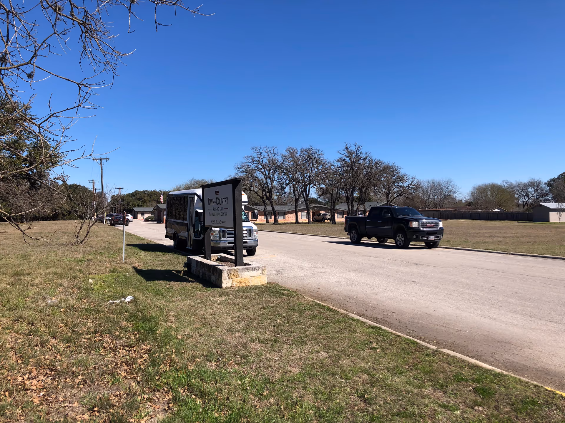 Wide outdoor street scene with a sign, grass verge, trees, and parked vehicles.