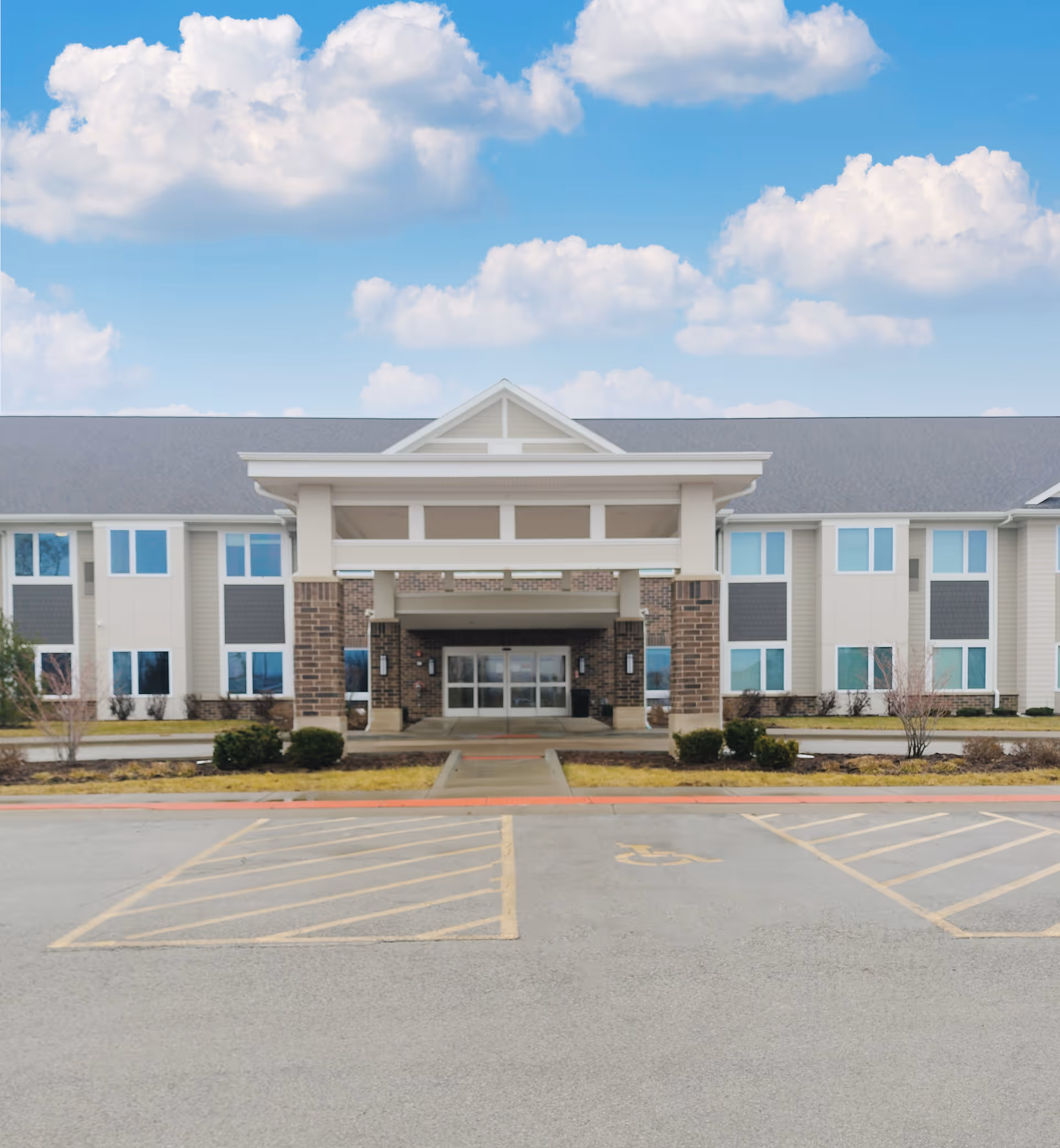 Front entrance of a two-story senior living building with a covered portico, parking spaces, and a blue sky with clouds.