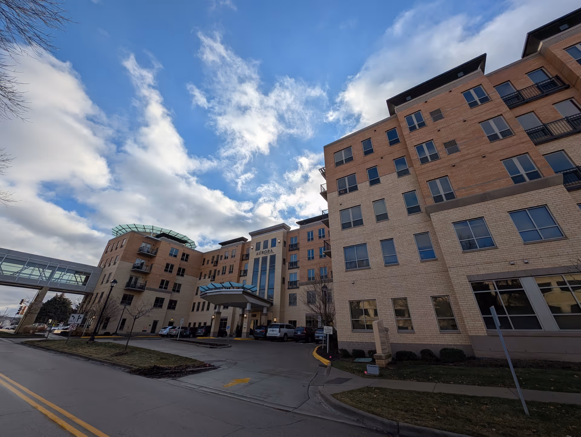 Exterior front view of a multi-story brick senior living building labeled "Aurora" with a covered entrance and skybridge.