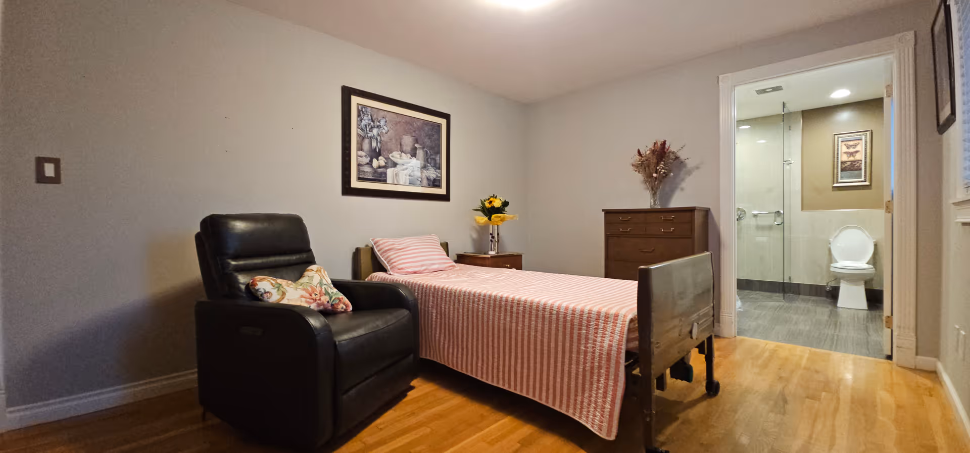 A bedroom featuring a bed, recliner, dresser, nightstand with flowers, and an open bathroom doorway.
