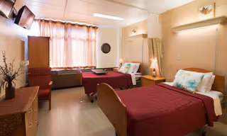 A well-lit bedroom with two single beds, each with a red bedspread and decorative pillows. There is a wooden nightstand with a lamp between the beds, a dresser with a vase and flowers, a wall-mounted TV, and a window with sheer curtains allowing natural light to enter the room.