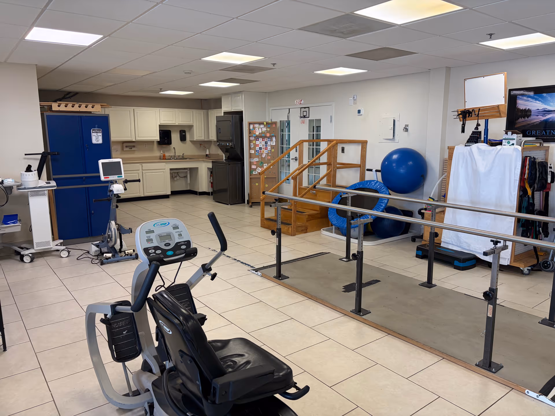 Interior rehab gym in a senior facility with exercise bike, parallel bars, and large blue ball.