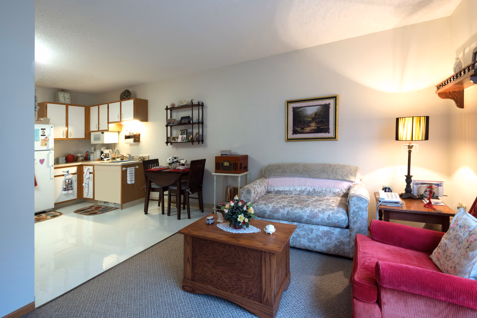 A cozy living area with a floral-patterned loveseat, a red armchair, and a wooden coffee table with a flower arrangement. To the left is a small dining table with four chairs and a compact kitchen featuring white cabinets, a refrigerator, microwave, and various kitchen items. The room is softly lit with a table lamp and wall-mounted light fixtures, and a framed landscape painting hangs on the wall above the loveseat.