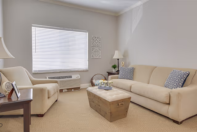 Well-lit living room with a beige sofa and armchair, a wooden ottoman coffee table, side tables with lamps, and a window with blinds.