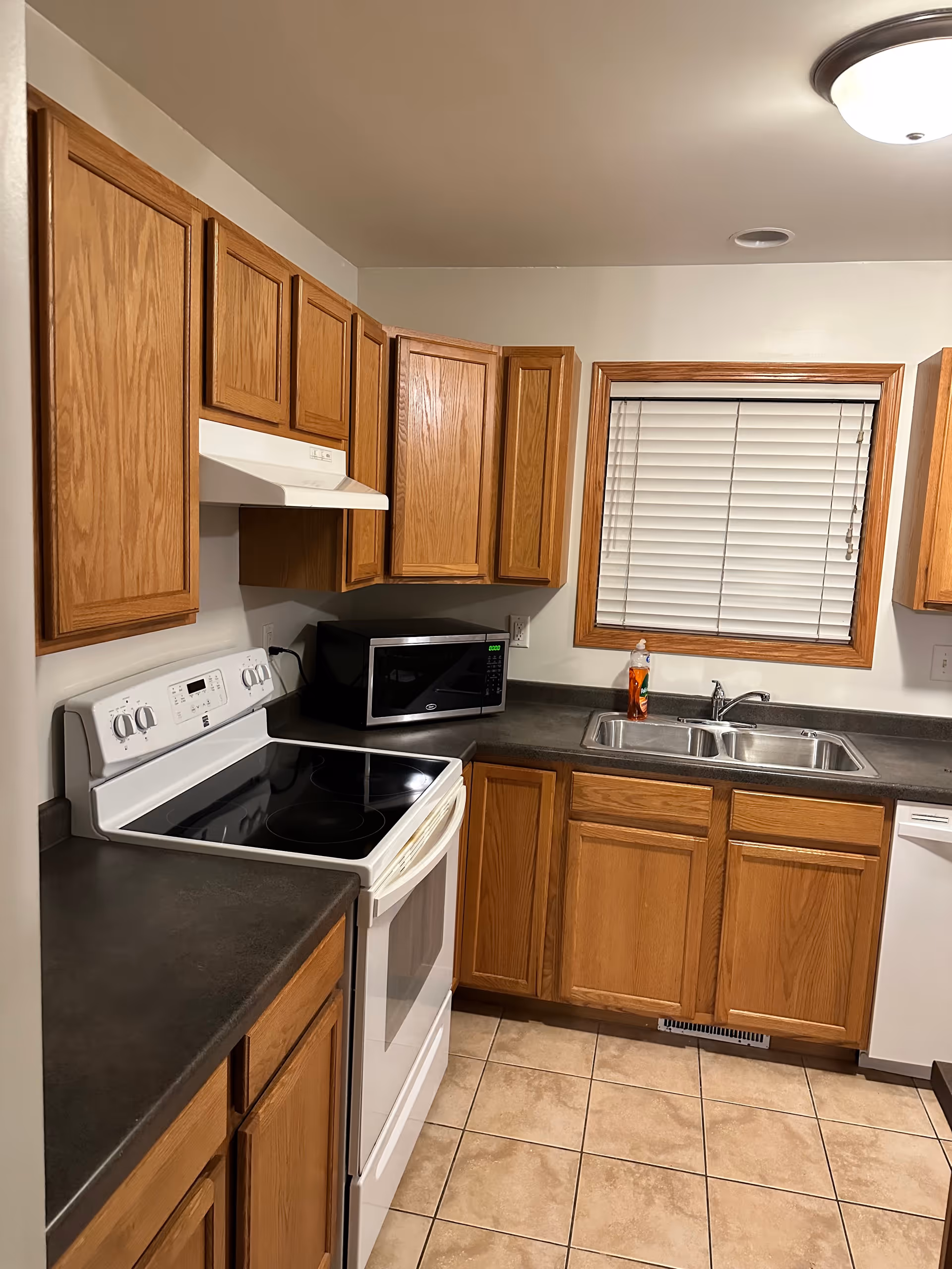 Compact kitchen with wooden cabinets, stove, microwave, sink, and tiled floor.