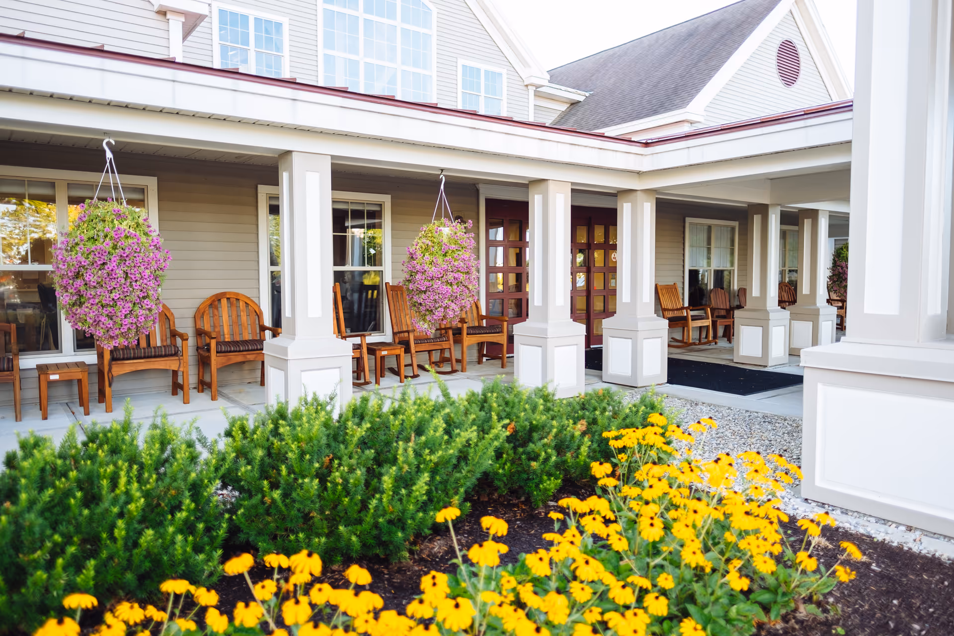 Front porch area of The Glen at Hiland Meadows with wooden chairs lined up along the wall under a covered walkway, hanging baskets of purple flowers, and a garden bed with yellow flowers and green shrubs in the foreground.