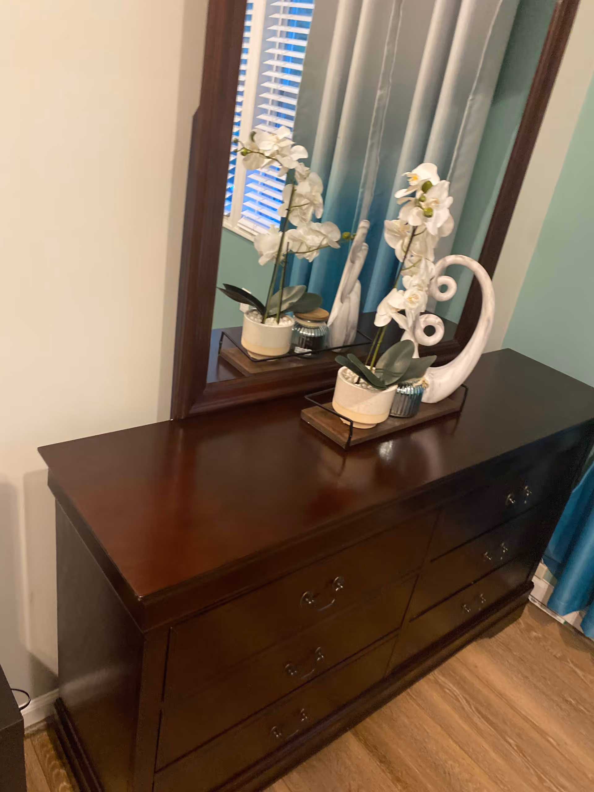 A dark wooden dresser with six drawers topped with a large mirror. On the dresser are two white orchid plants in pots and a white decorative sculpture. The background shows a window with white blinds and blue curtains.