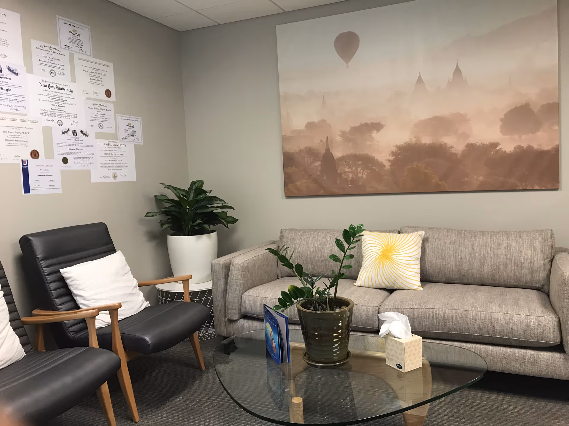 Cozy waiting area with grey sofa, two black chairs, glass table, plants, and wall art.